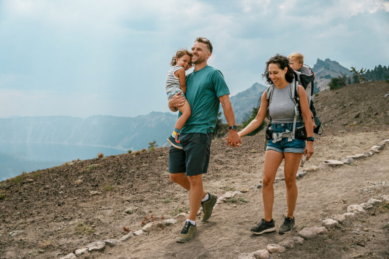 A fit young dad carries his three year old daughter and holds hand with his multiracial wife who is carrying their one year old son in a backpack baby carrier during a hike overlooking Crater Lake in Oregon.